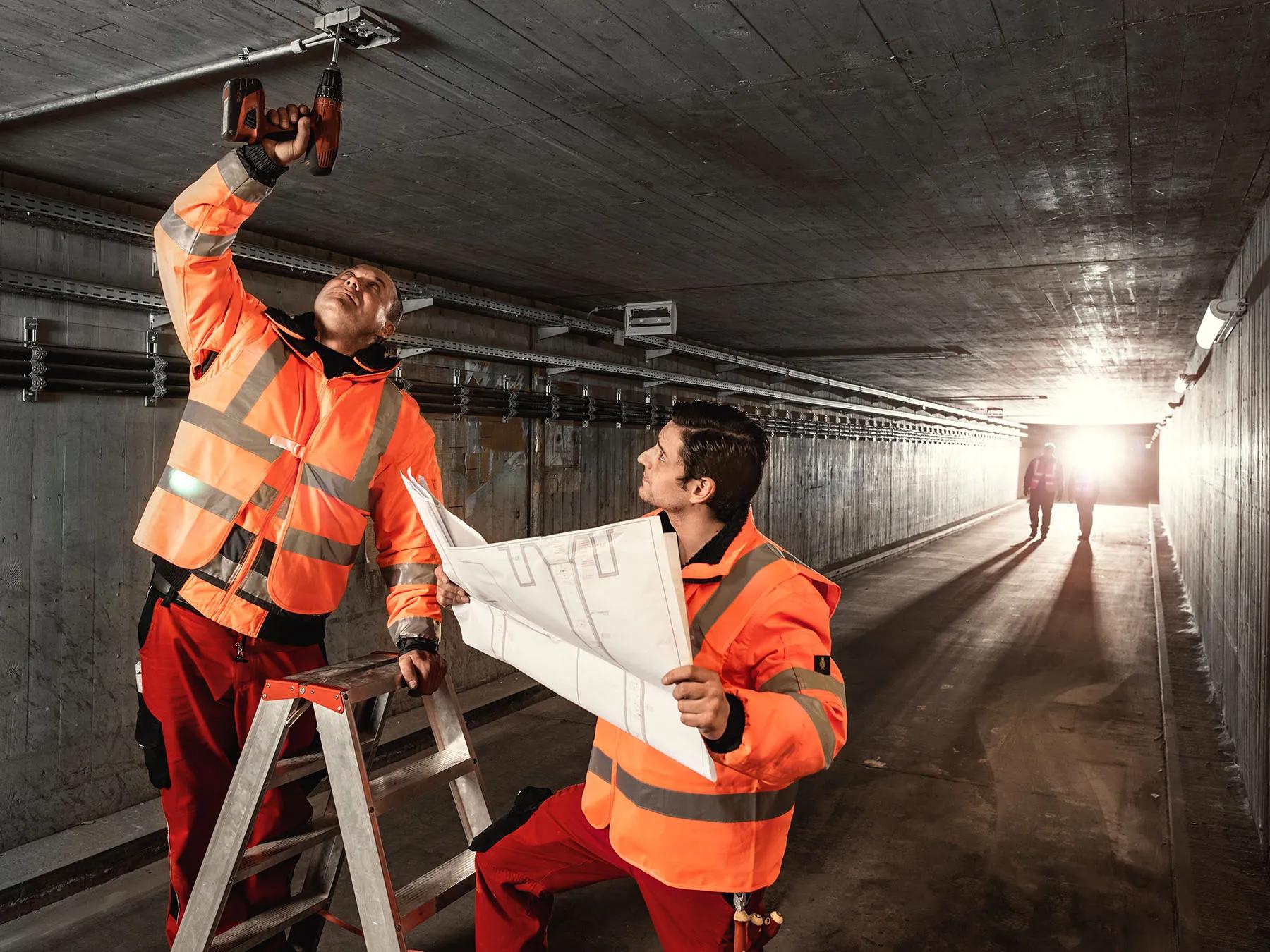 Ein Mitarbeiter arbeitet an der Decke eines Tunnels, ein weiterer steht mit einer Karte neben ihm, im Hintergrund sind zwei Mitarbeiter erkennbar
