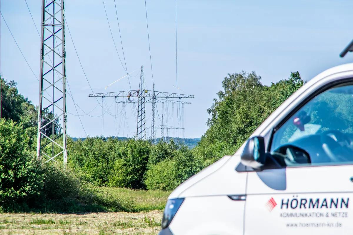 Bild von Freileitungsmasten, im Vordergrund ein Transporter mit der Aufschrift "HÖRMANN Kommunikation & Netze"